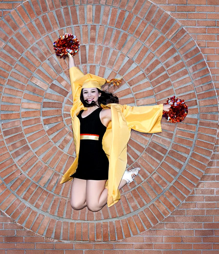 High school senior graduation portrait. A young woman in a black outfit, yellow graduation gown and cap, white sneakers, holding red and silver pom-poms, jumping in front of a circular brick wall design, smiling happily.