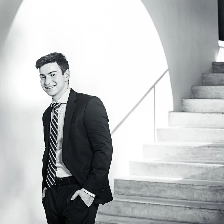 Brophy Phoenix Senior. A young man in a black suit, white shirt, and striped tie standing next to a staircase in a modern, minimalist interior.