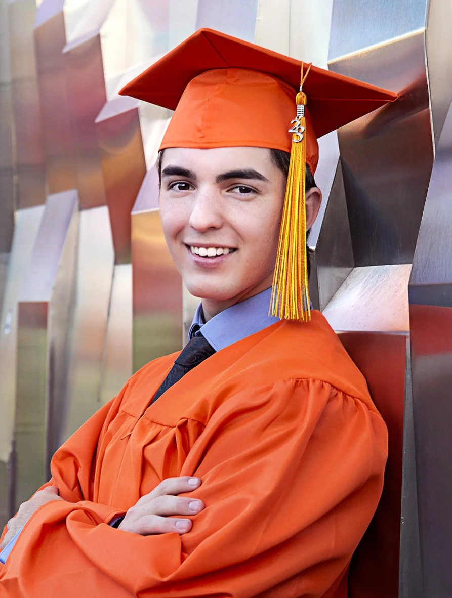 Senior young man in orange High school graduation cap and gown smiling, standing with arms crossed, in front of metallic geometric wall. Senior photography in Scottsdale.