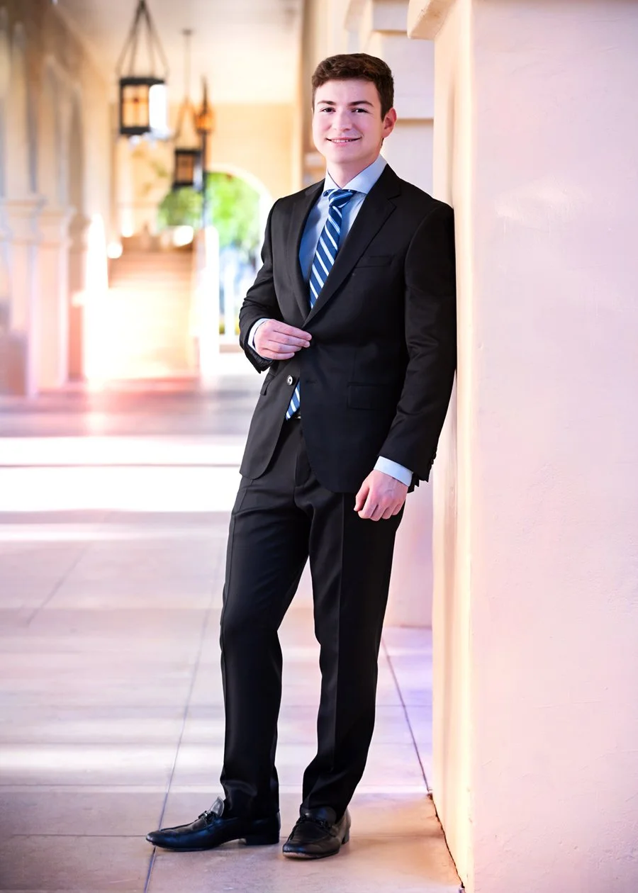 A young man in a black suit and blue striped tie standing and leaning against a wall in a well-lit corridor, smiling at the camera. High school senior photos in Scottsdale Arizona.