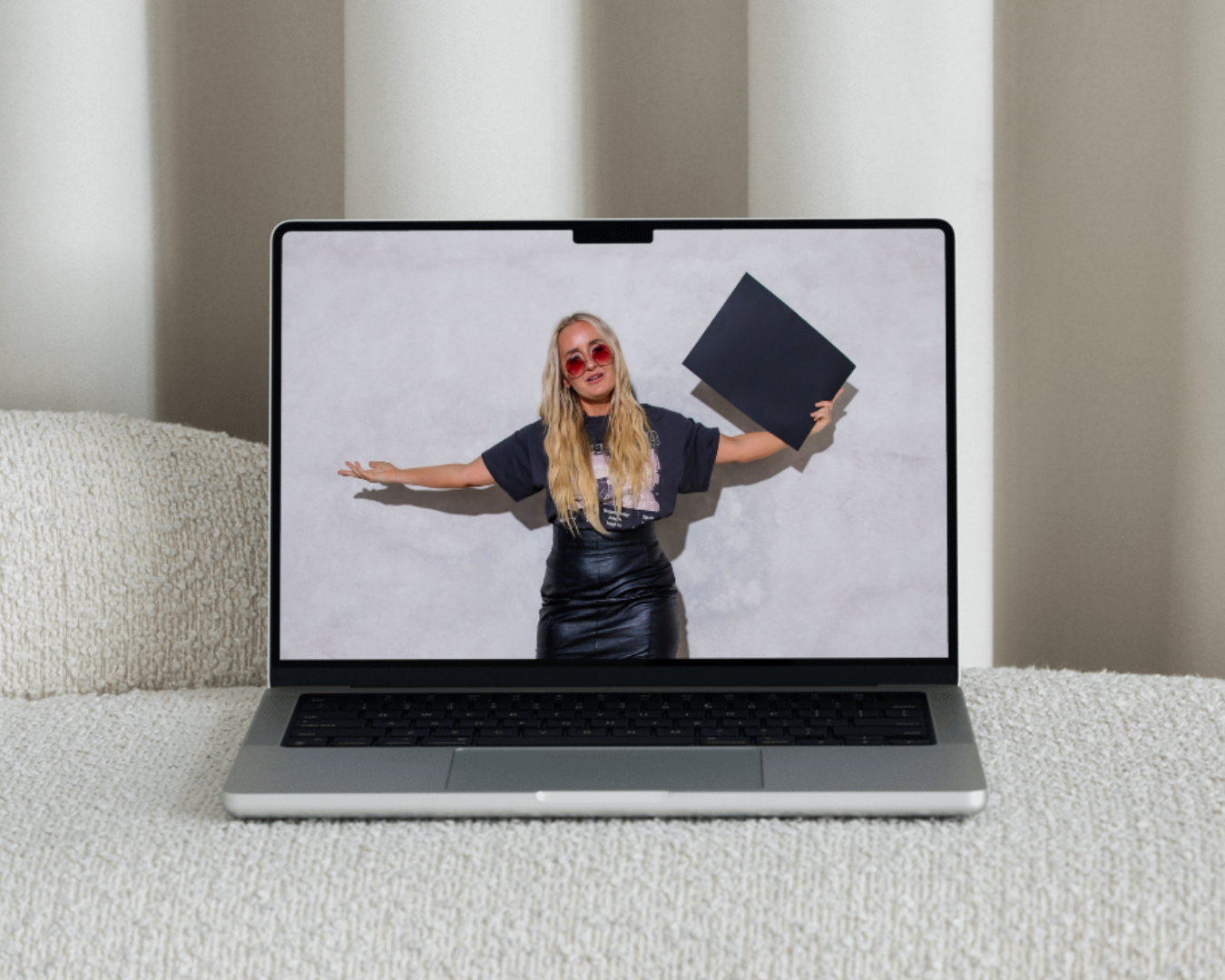 Laptop displaying an image of a woman with long hair, wearing sunglasses, black outfit, and holding a large black square object, standing against a light gray background, laptop on a textured fabric surface.