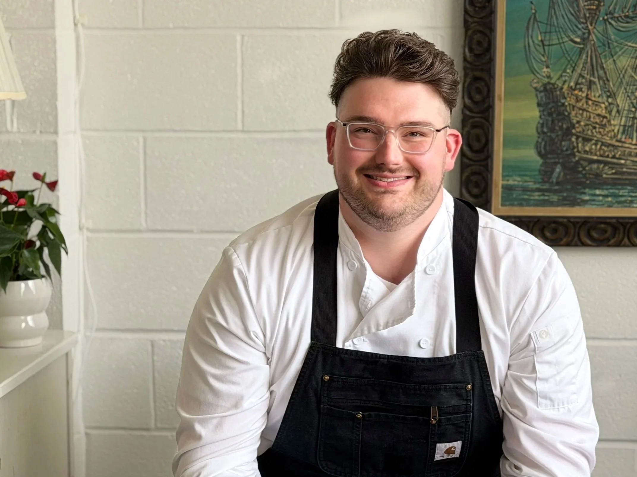 Man in a black chef coat standing indoors with a framed painting on the wall.