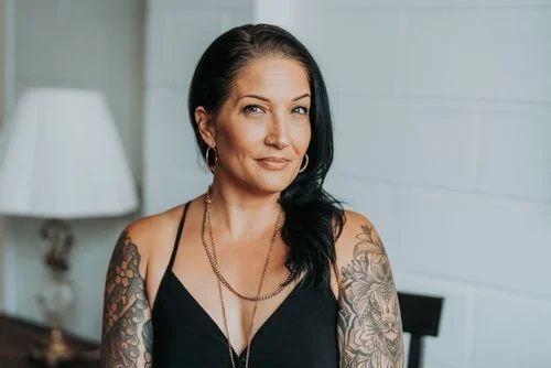 Woman with tattoos wearing a black top and necklace, sitting indoors with a lamp in the background.