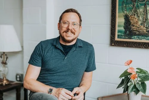 A man wearing glasses and a blue polo shirt sitting in a room with a lamp and a plant, smiling at the camera.