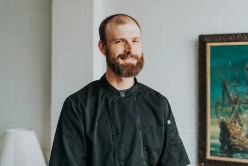 Man in a black chef coat standing indoors with a framed painting on the wall.