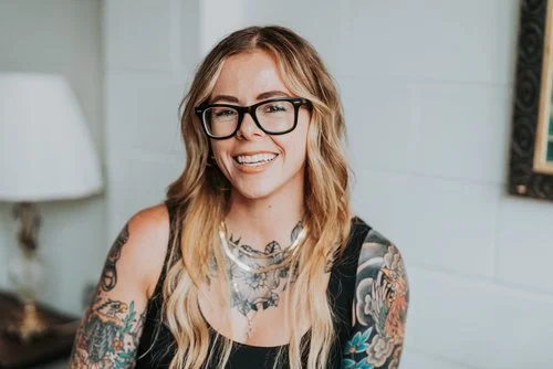 Smiling woman with tattoos and glasses in a room with a lamp.