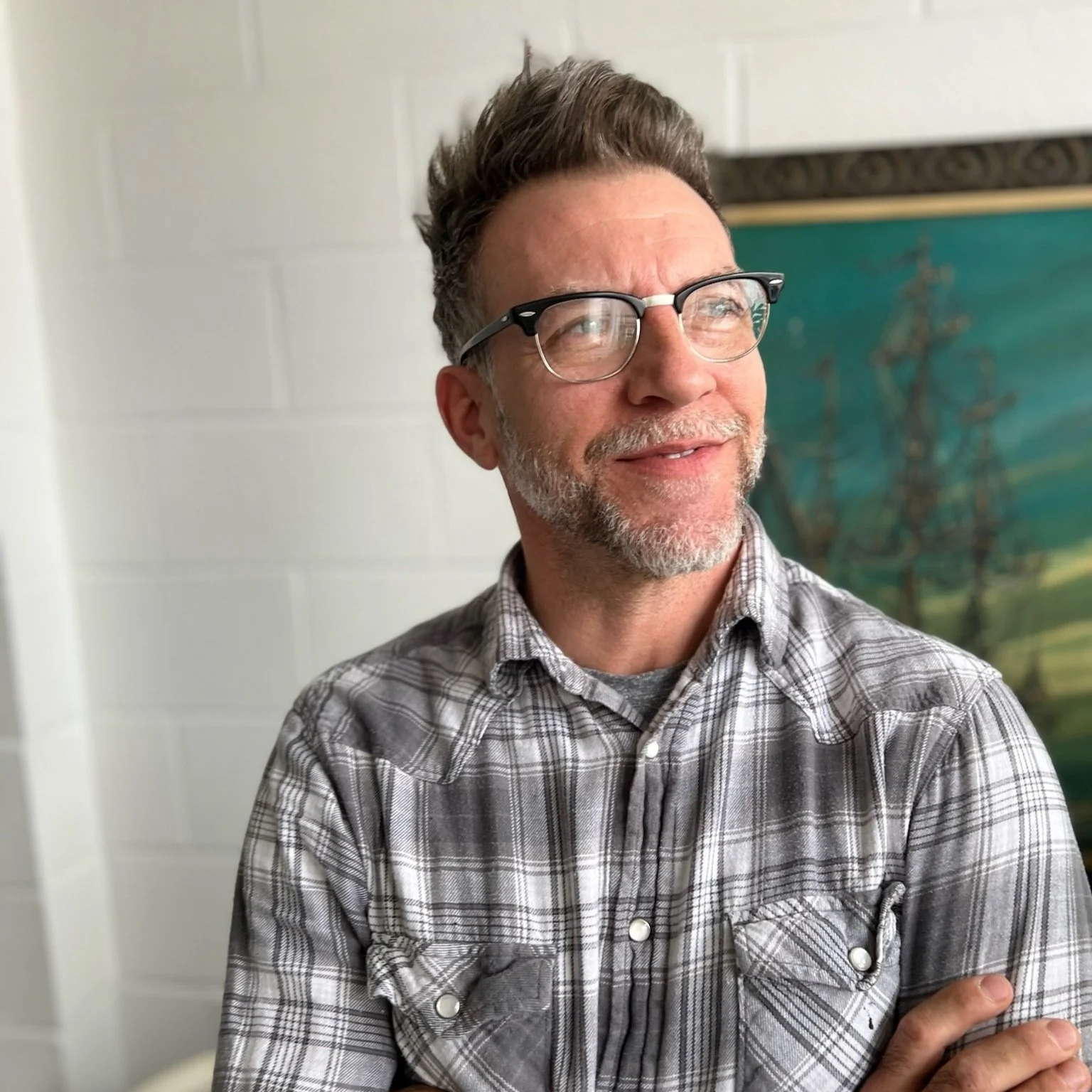 A man with glasses and a plaid shirt looking to the side, standing indoors.