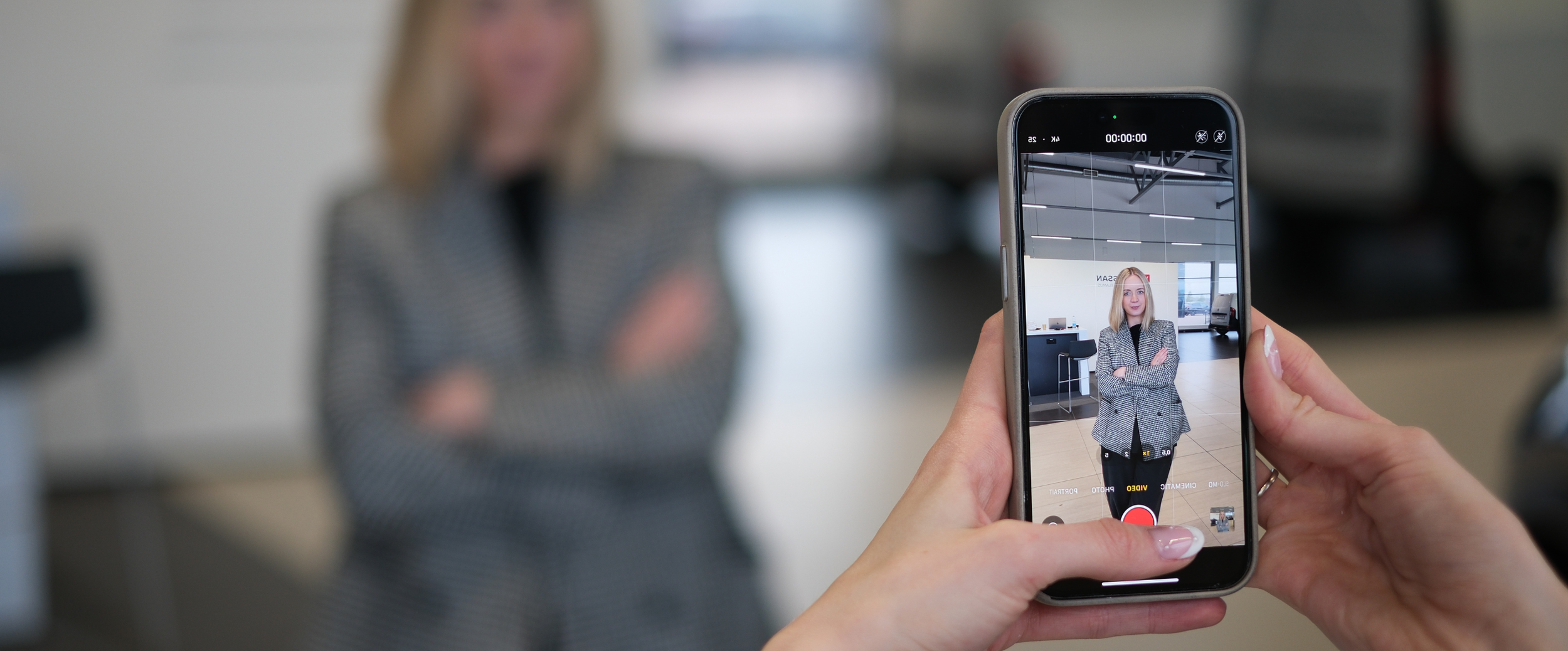 Person taking a photograph of a woman standing in a modern office with a smartphone.