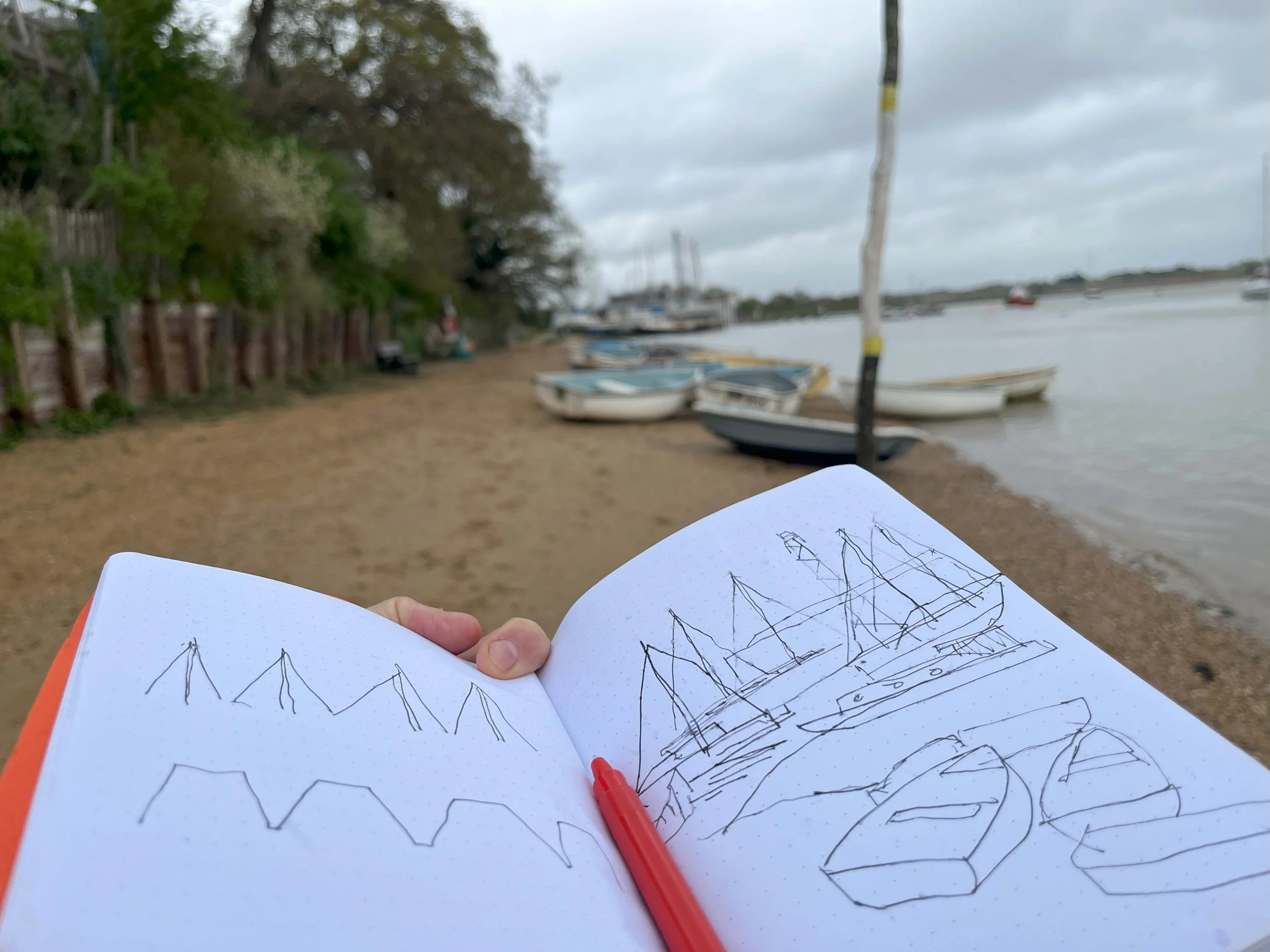 Architect’s sketchbook held up on Waldringfield quay, showing drawings of tapered roof forms and boat masts with boats on the shoreline in the background