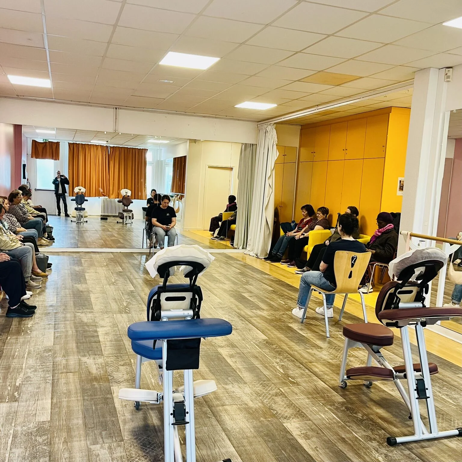 Salle de physiothérapie avec des patients assis en cercle, plusieurs fauteuils et équipements médicaux.