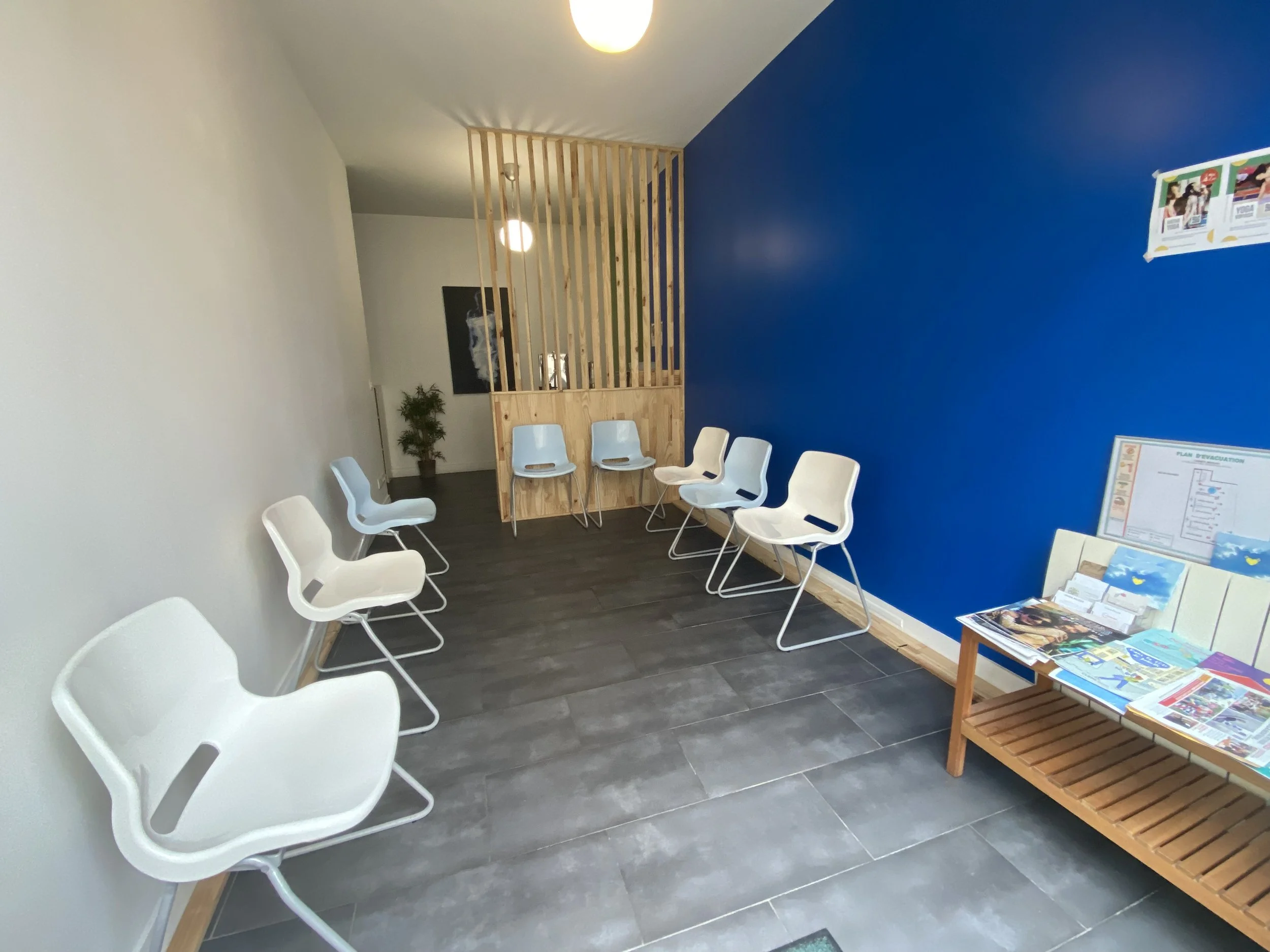 Salle d'attente avec neuf chaises blanches disposées contre les murs blanc et bleu. Il y a un banc en bois avec des magazines dessus, une petite plante verte et un panneau d'information.