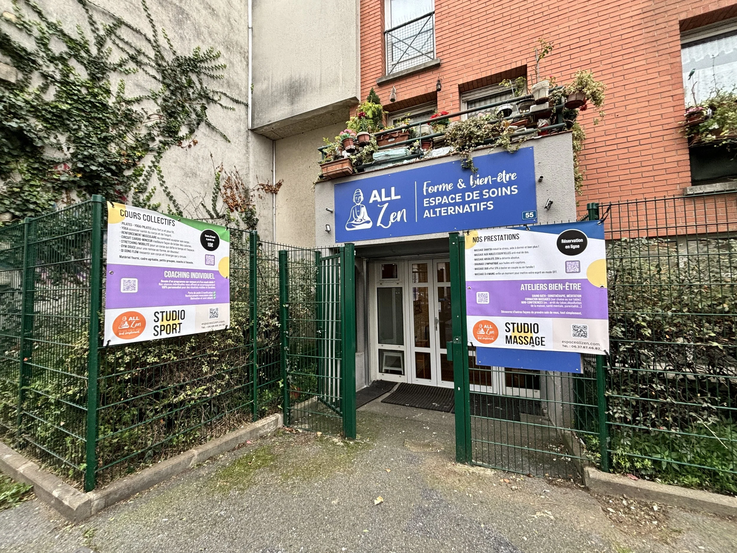 Entrée d'un centre de bien-être appelé 'ALL Zen', avec une porte blanche encadrée par une clôture verte. Au-dessus de l'entrée, un grand panneau bleu indique le nom et la nature de l'établissement. Deux affiches promotionnelles sont fixées à la clôtu