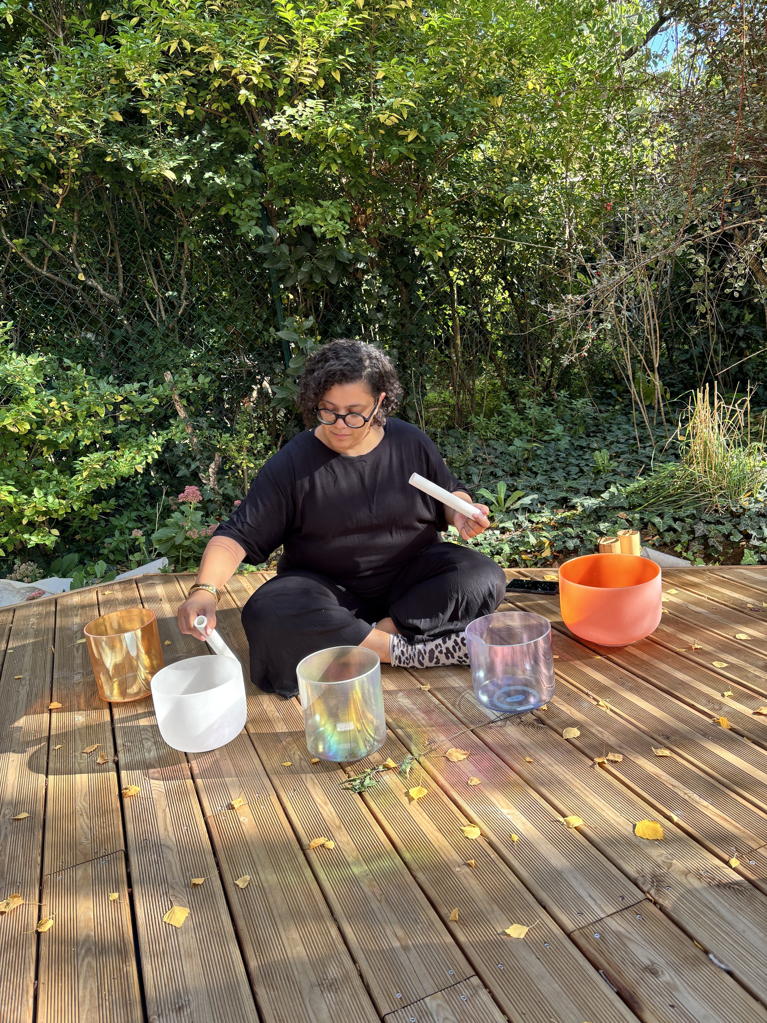 Une femme assise sur une terrasse en bois, jouant avec des bols en verre de différentes couleurs, dans un environnement extérieur verdoyant.