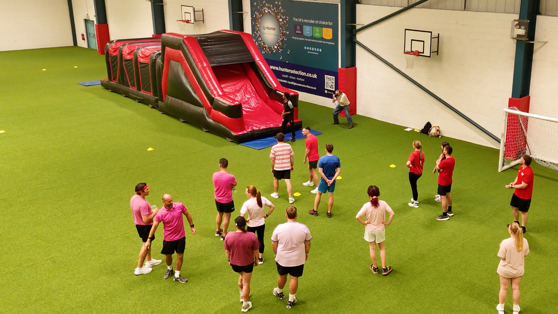 Indoor sports facility with a green turf field where people are gathered for a training session or event. There is an inflatable obstacle course set up on the field, and some participants are standing in a line or talking. Branded banners and a baske