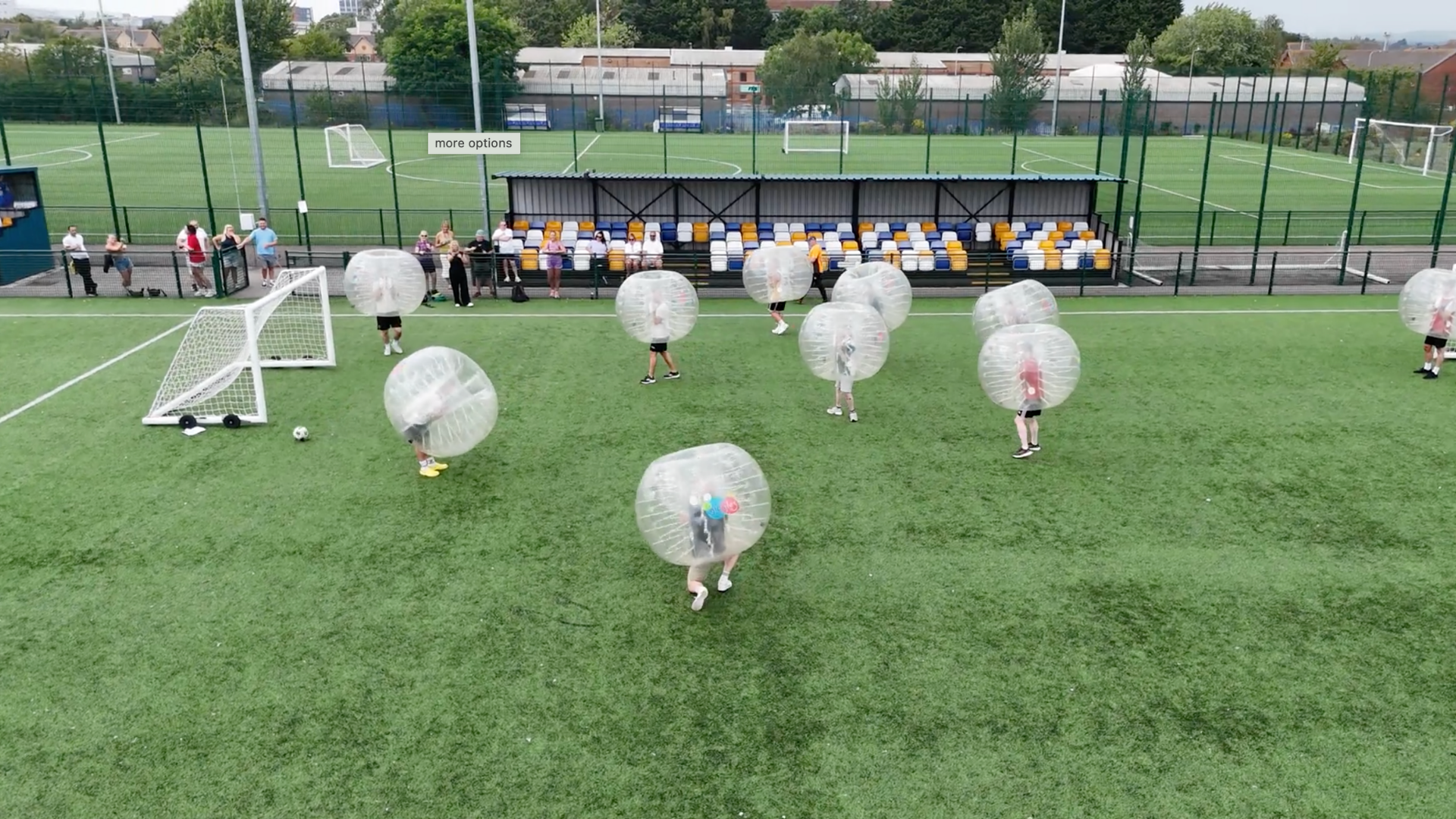 People engaging in a bubble soccer game on a green sports field, with some spectators sitting on a covered bench area and others standing behind a fence, in a sports complex surrounded by trees and buildings.