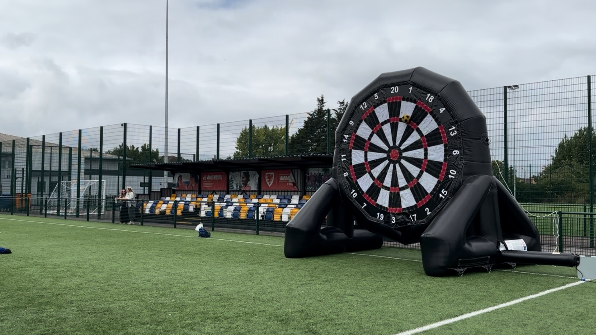 Inflatable dartboard set on a sports field with stadium seating and a fence in the background, cloudy sky overhead.