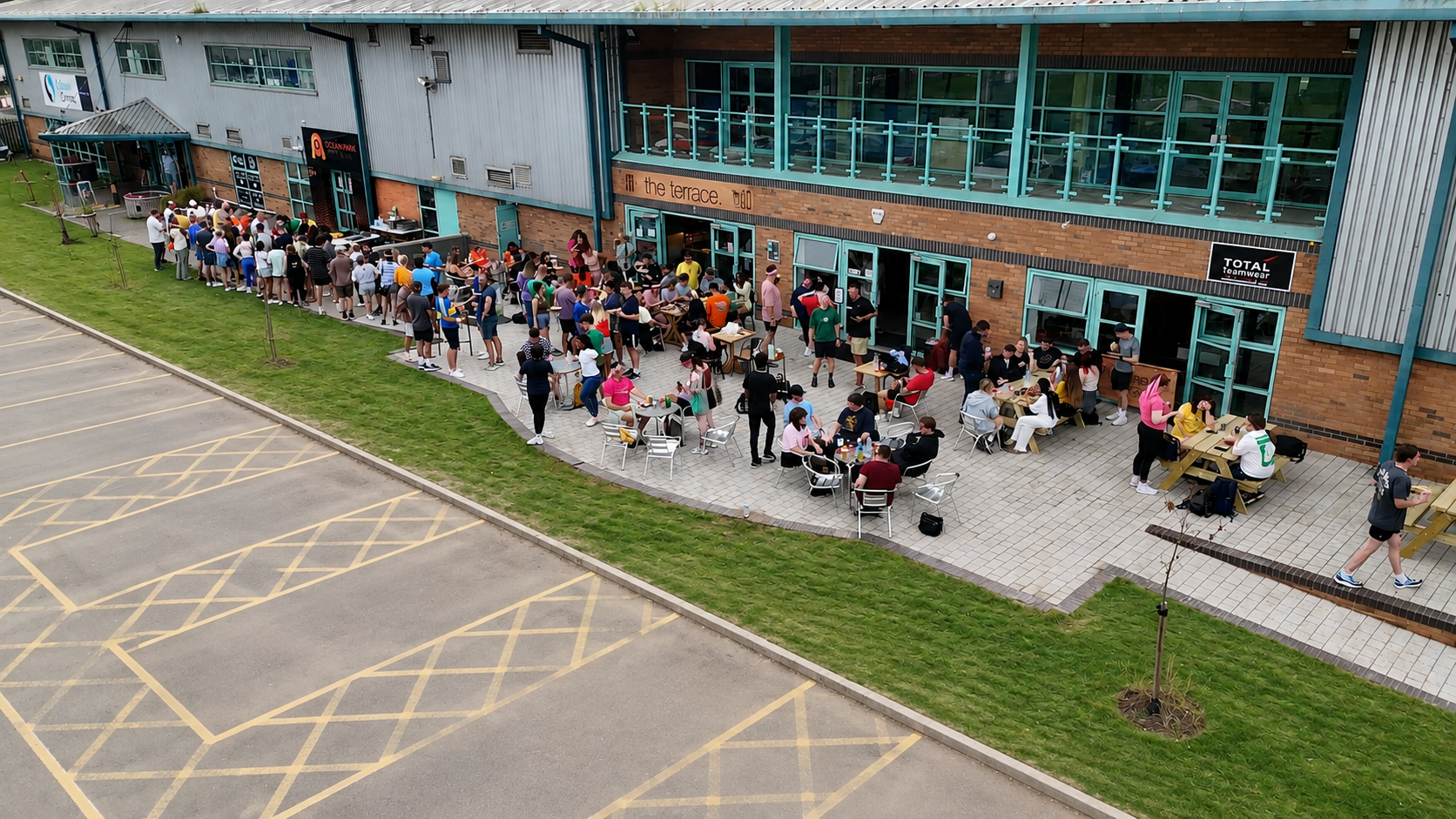 A large crowd of people standing in line outside a modern building with glass windows and brick walls, some seated at outdoor tables, in what appears to be a busy café or restaurant patio. Empty parking lot in foreground.