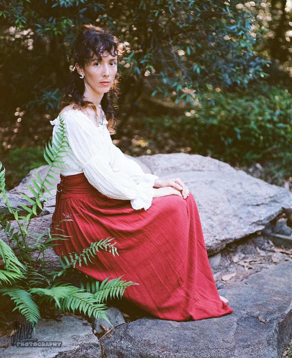 Pretty young lady in boho outfit sitting on rock in bushland environment