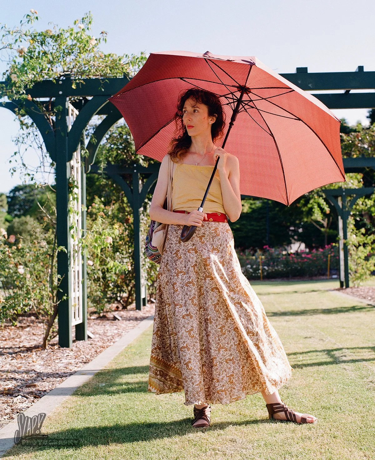 Pretty young lady with red umbrella posing amongst rose arches