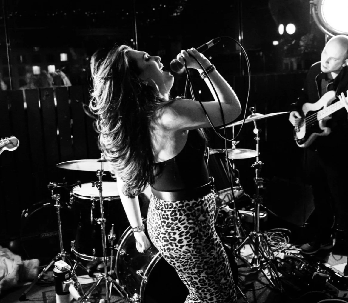 A woman singing passionately into a microphone on stage, with a drummer and guitarist in the background, in a black-and-white photo.