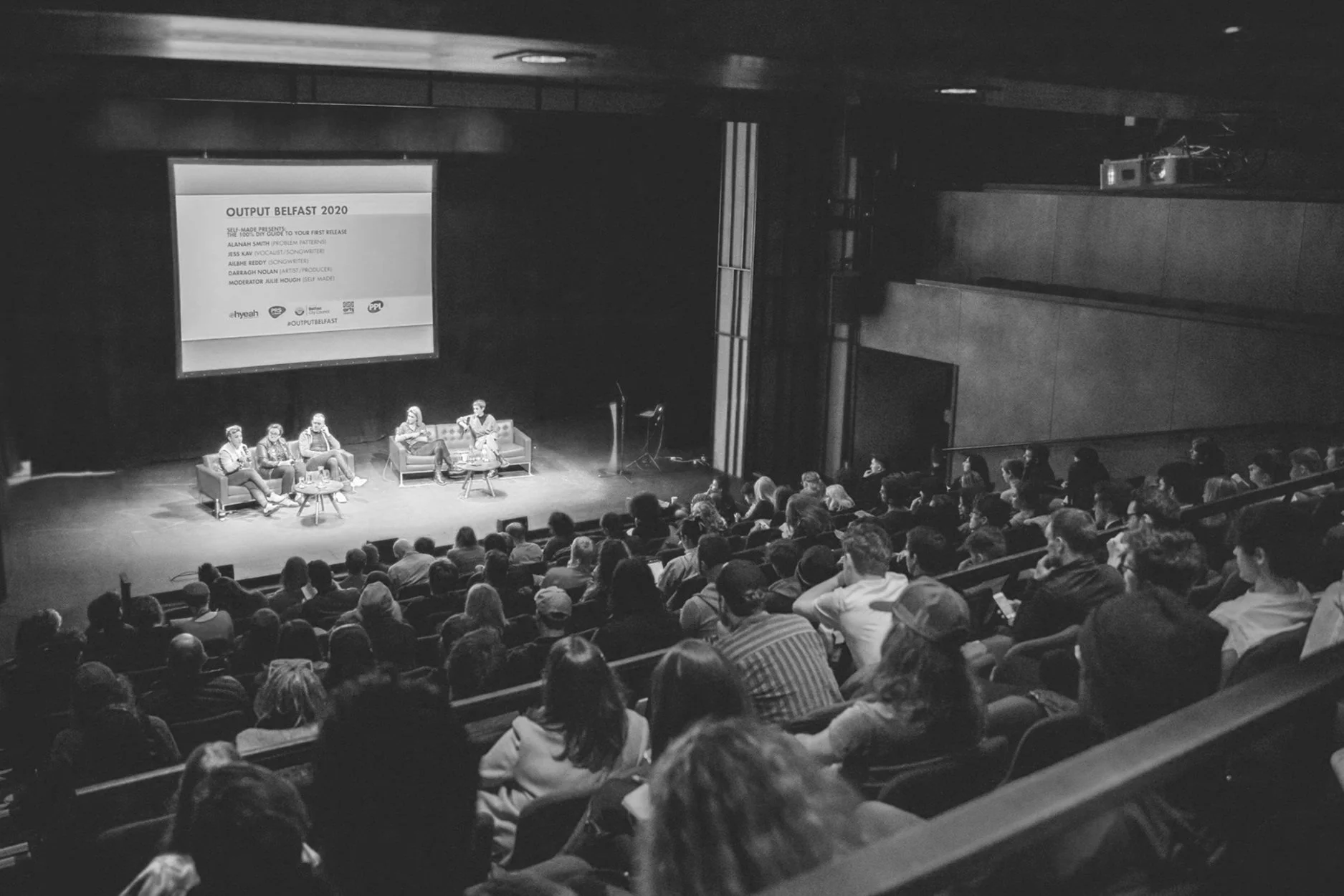 Black and white photo of a panel discussion in an auditorium. Five people sitting on stage with a large screen behind them displaying