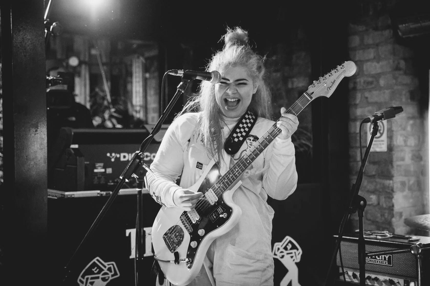 A woman passionately singing and playing an electric guitar on stage with a brick wall background, black and white photo.
