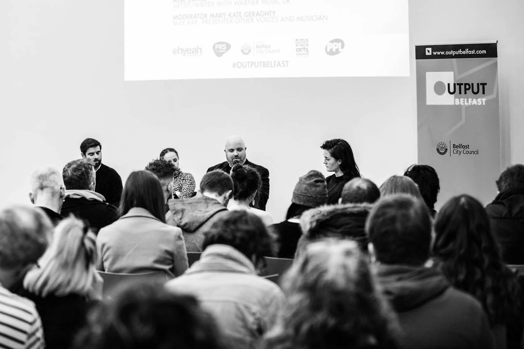 Panel of five speakers seated at a table at a Belfast event, with an audience in front of them and a screen above displaying logos and text.