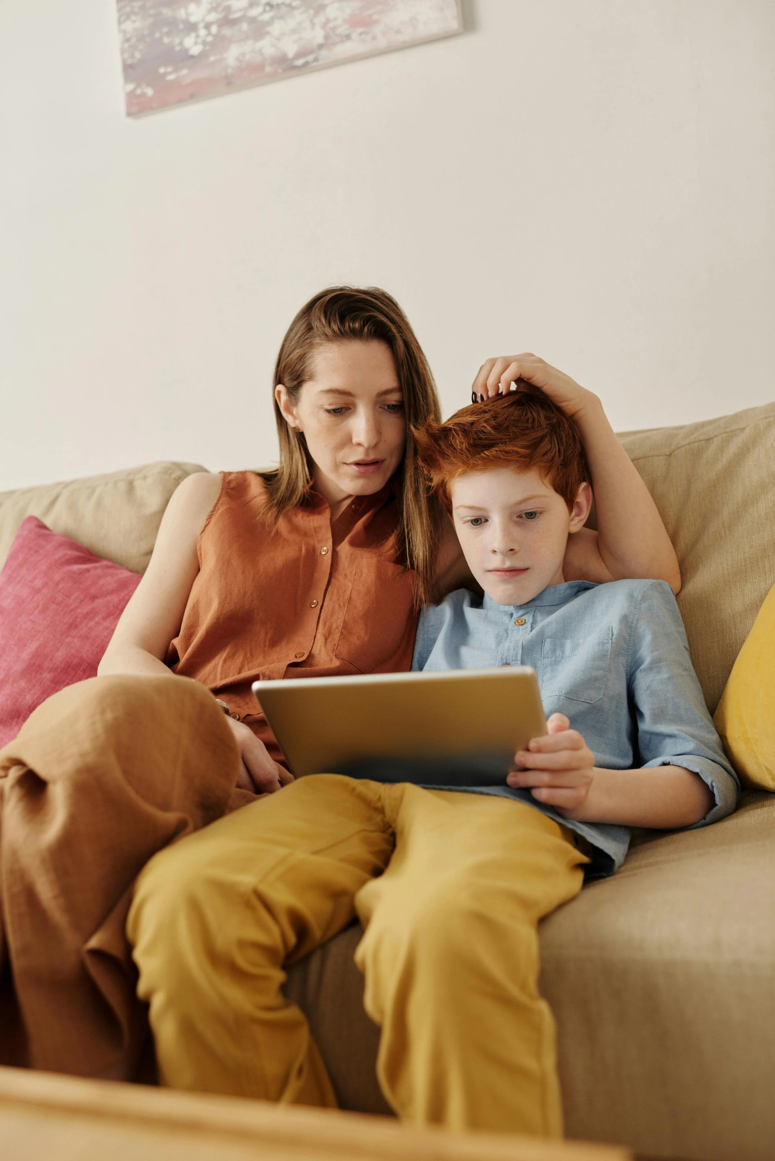 A woman and a young boy sitting on a beige couch, looking at a tablet. The woman is touching the boy's head, and they appear focused on the screen.