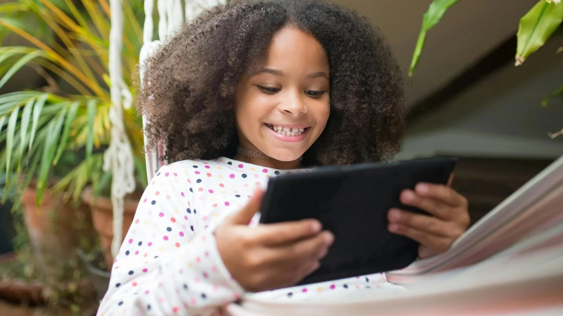 Young girl with curly hair smiling while looking at a tablet, sitting outdoors near green plants.