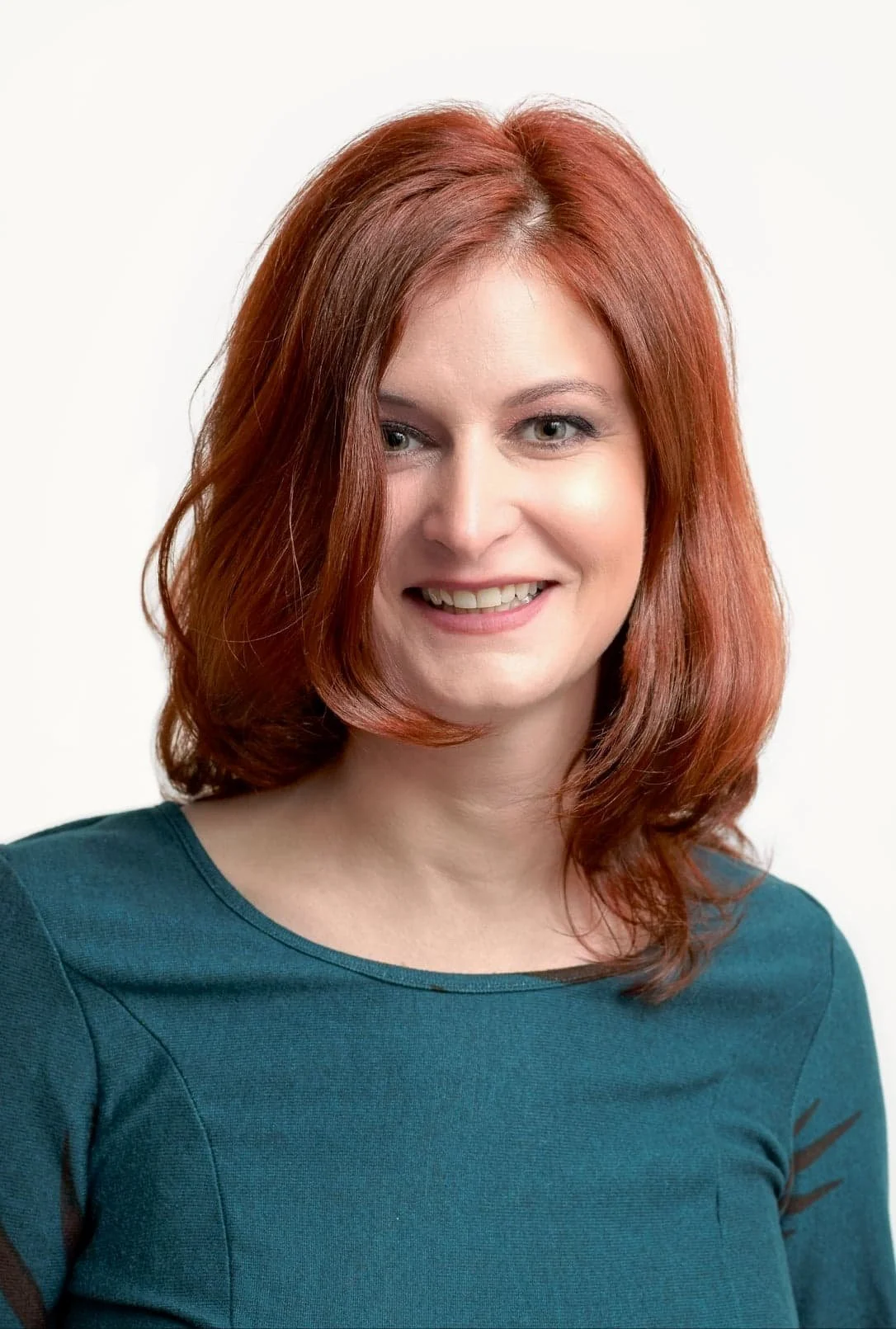Portrait of a woman with shoulder-length red hair, wearing a teal top, smiling and looking to the side against a plain white background.
