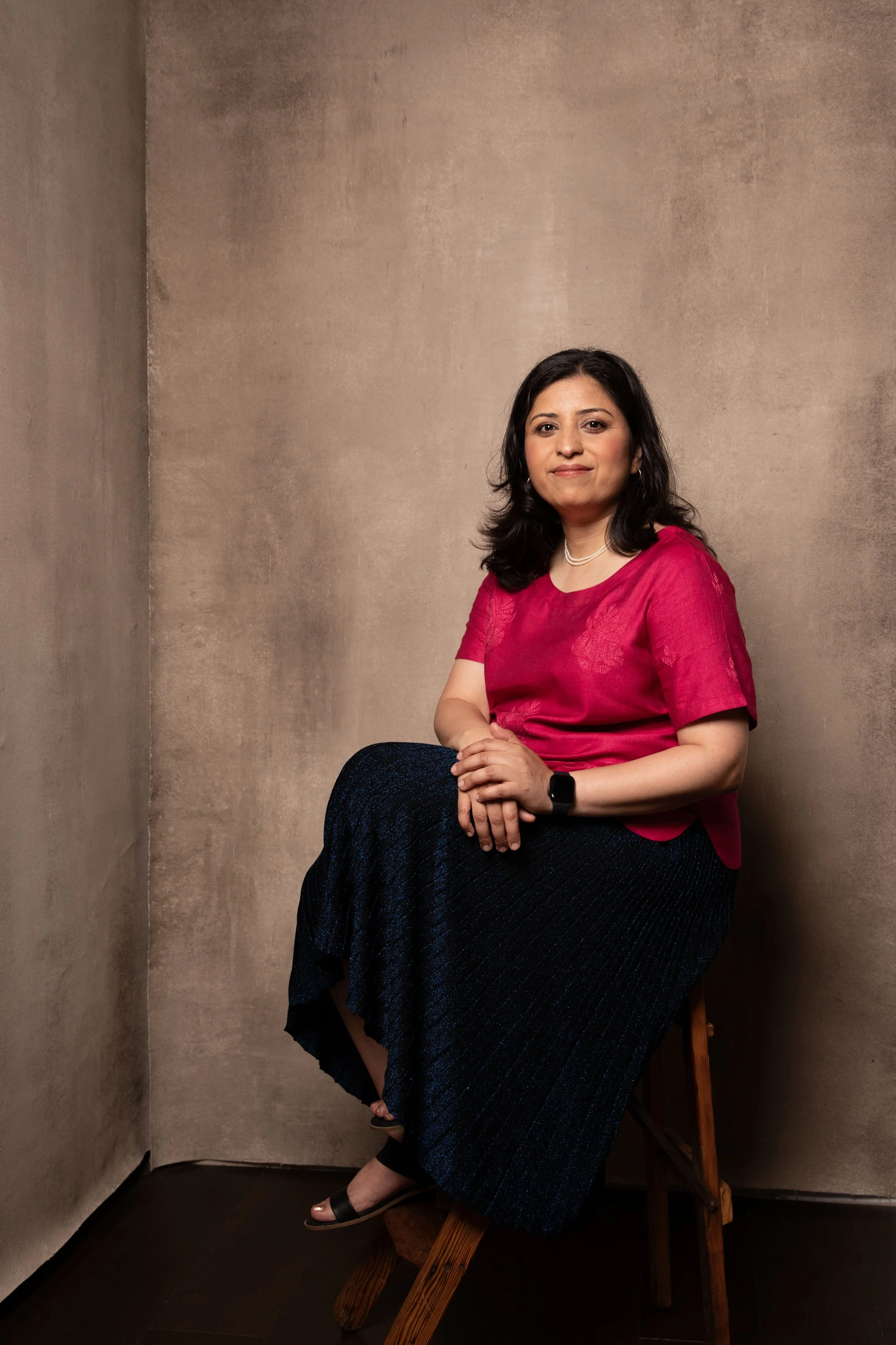 A woman with dark hair in a red blouse sitting on a wooden chair, with a neutral expression, against a plain textured wall.