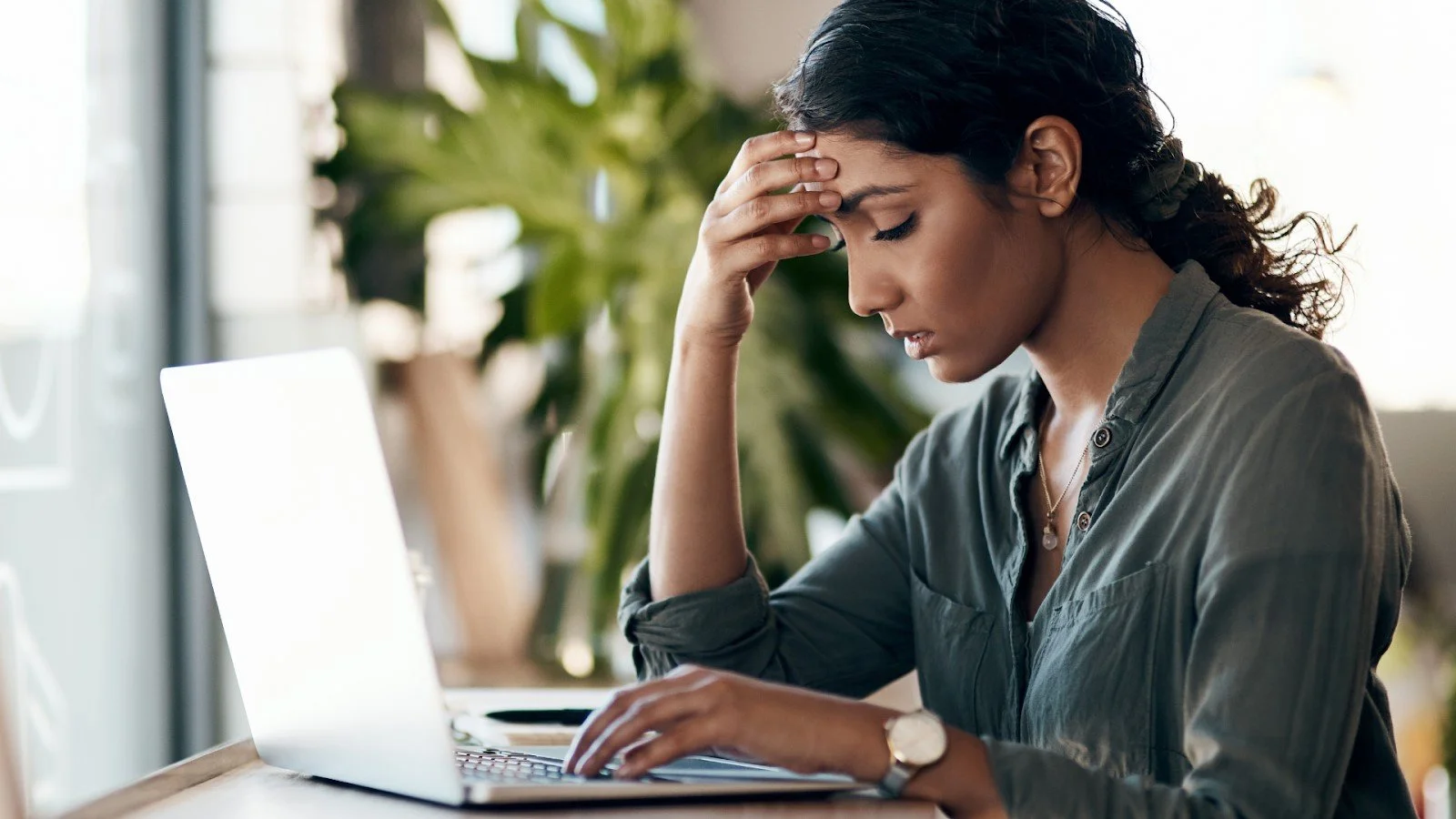 A woman sitting at a desk with a laptop, holding her forehead with a stressed expression.
