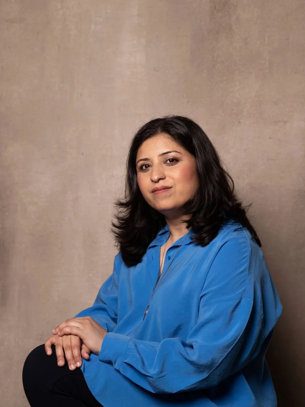 A woman with shoulder-length dark hair, wearing a blue blouse, sitting against a plain beige background, looking confidently at the camera.