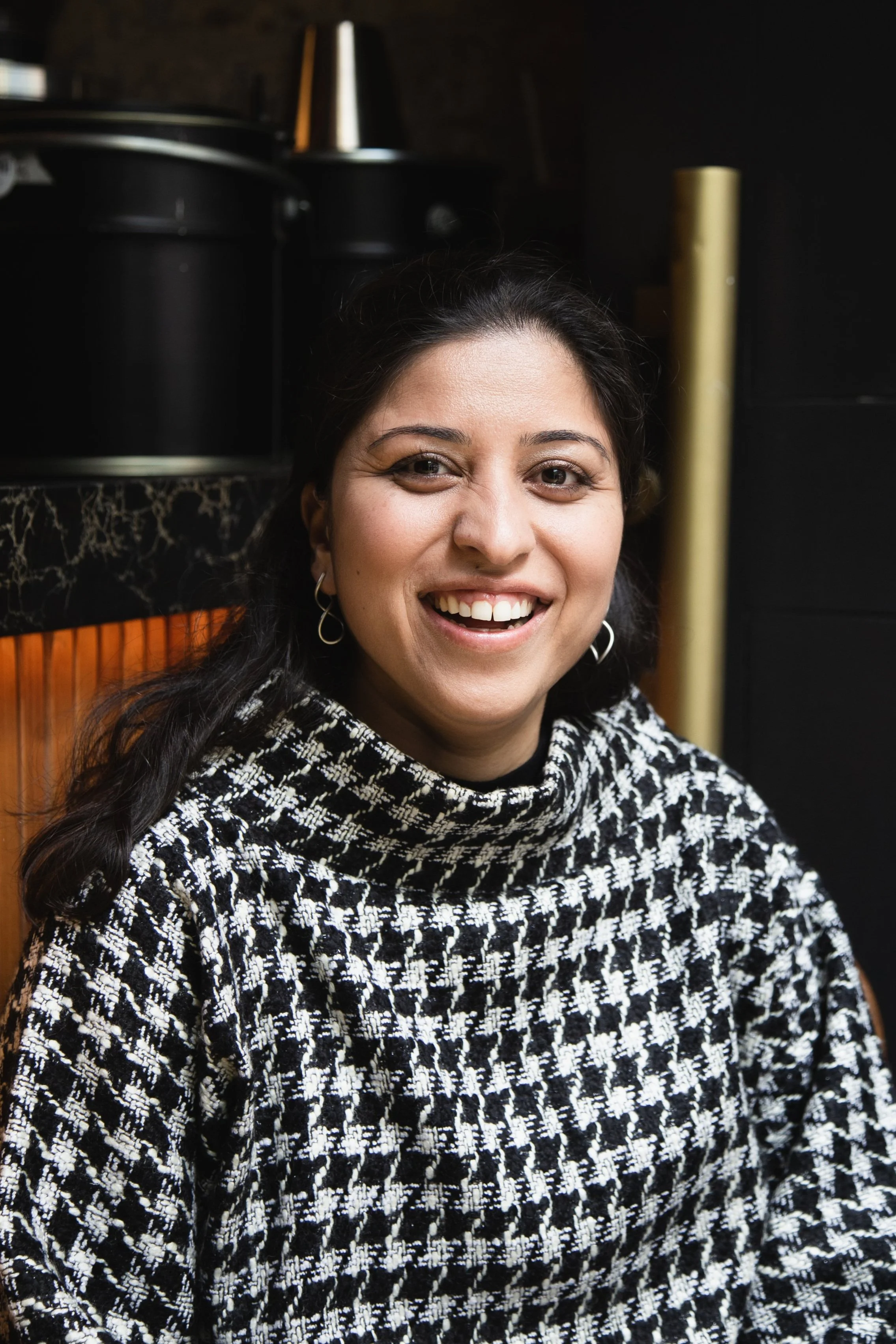 Close-up of a smiling woman with dark hair, wearing a black and white patterned turtleneck sweater, sitting indoors with a dark background.