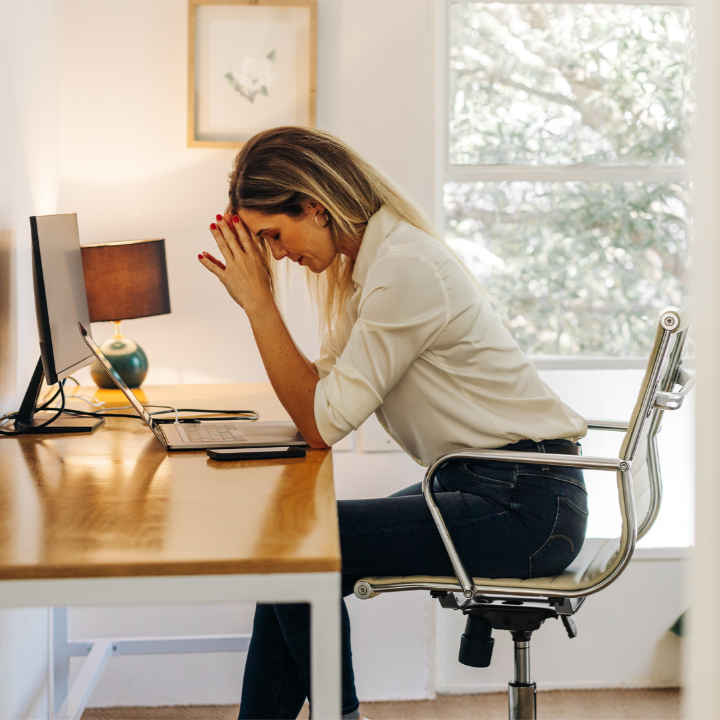 Frustrated woman sitting at a desk with a computer, holding her head in her hands.