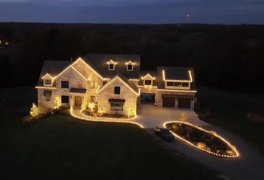 Aerial view of a large home at night with warm white Christmas lights on the roofline and driveway