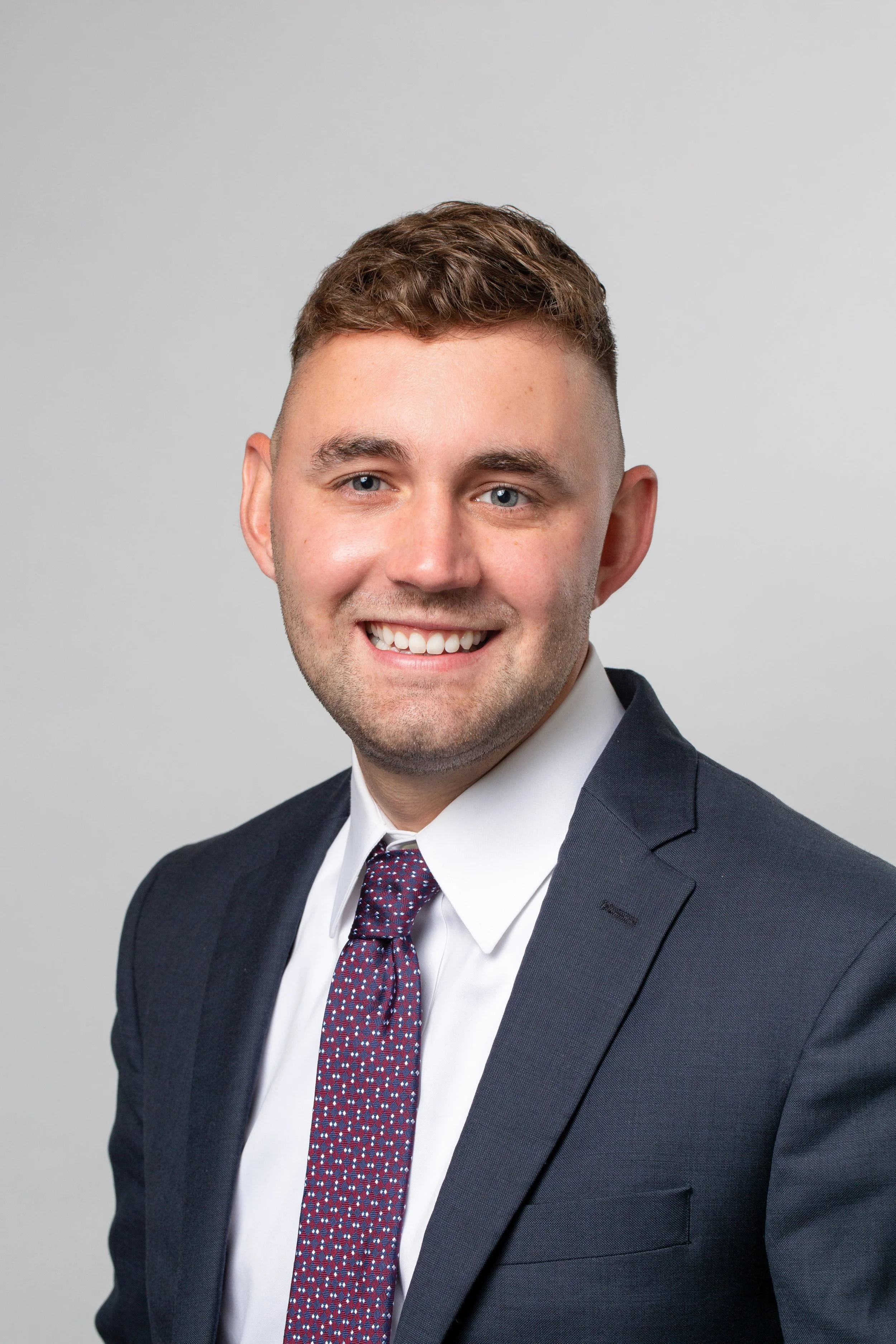 Headshot of a young man with brown hair, blue eyes, smiling, wearing a dark blue suit, white dress shirt, and patterned tie.