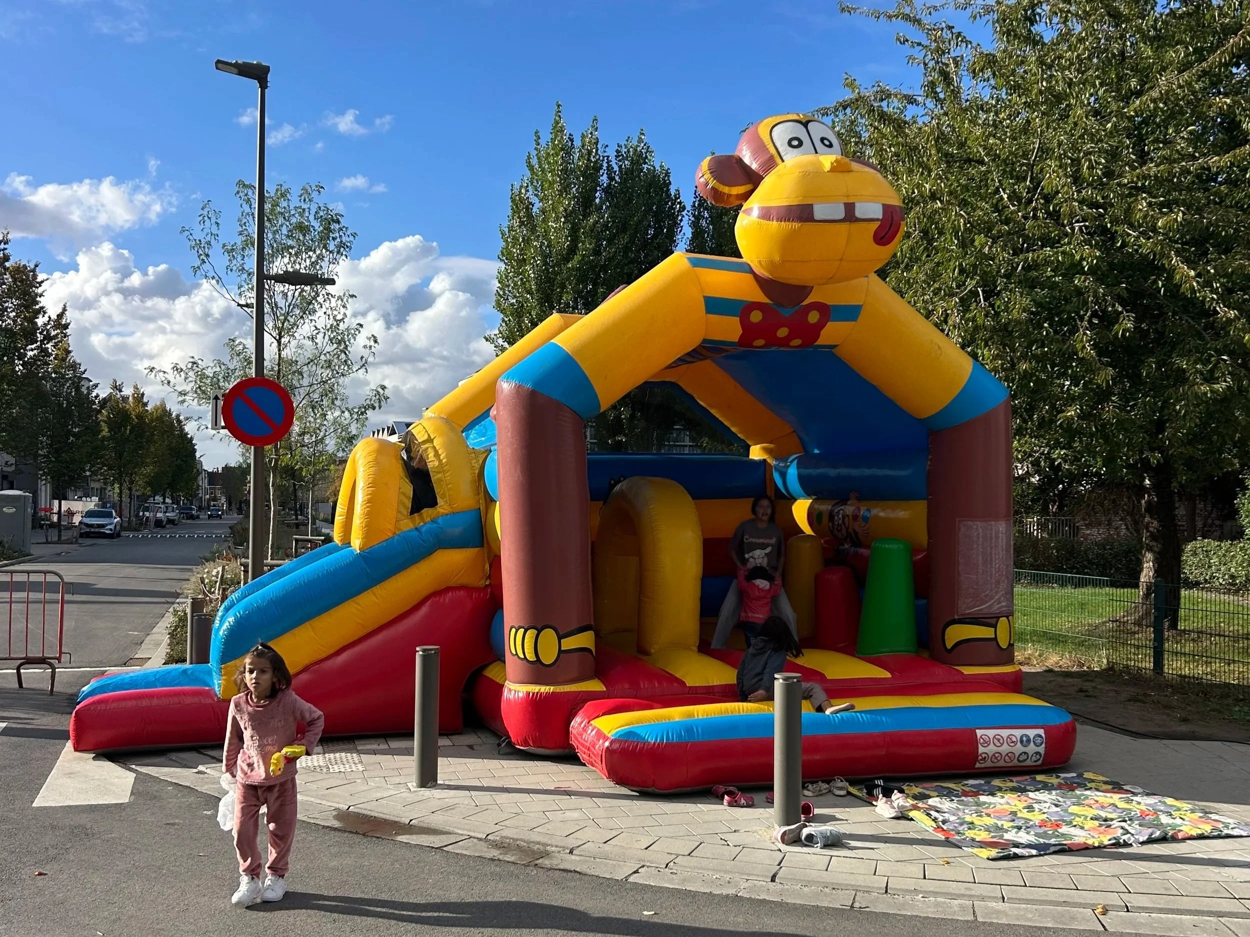 Een kleurrijke opblaasbare glijbaan met cartoon karakter bovenop, met kinderen die erop spelen en eromheen. Gelegen op een trottoir in een stadswijk, met bomen, een park en een blauwe lucht met wolken op de achtergrond.