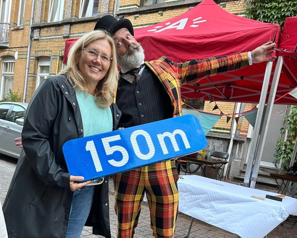Een vrouw en een man poseren voor een straatmarkt. De vrouw houdt een bord vast met de tekst '150 m'. De man draagt een kleurrijk kostuum en een zwarte hoed.