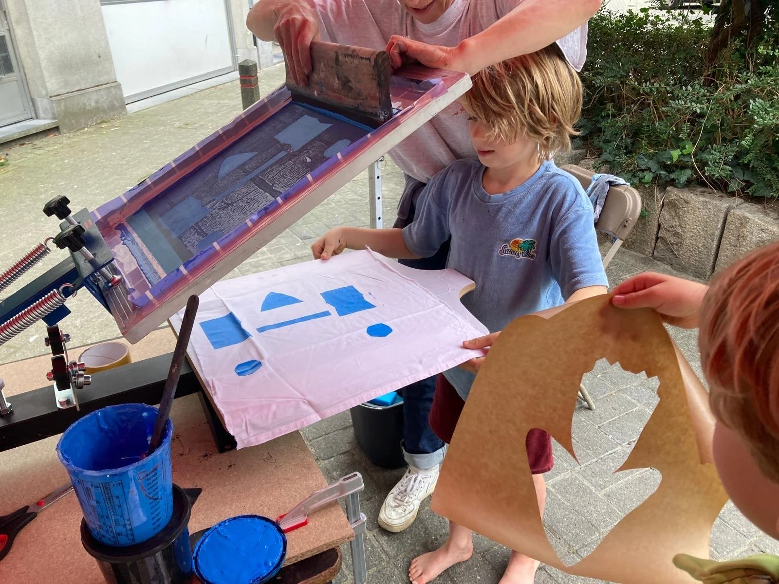Twee kinderen en een volwassene maken zeefdrukken met blauw inkt op een stuk textiel buiten.