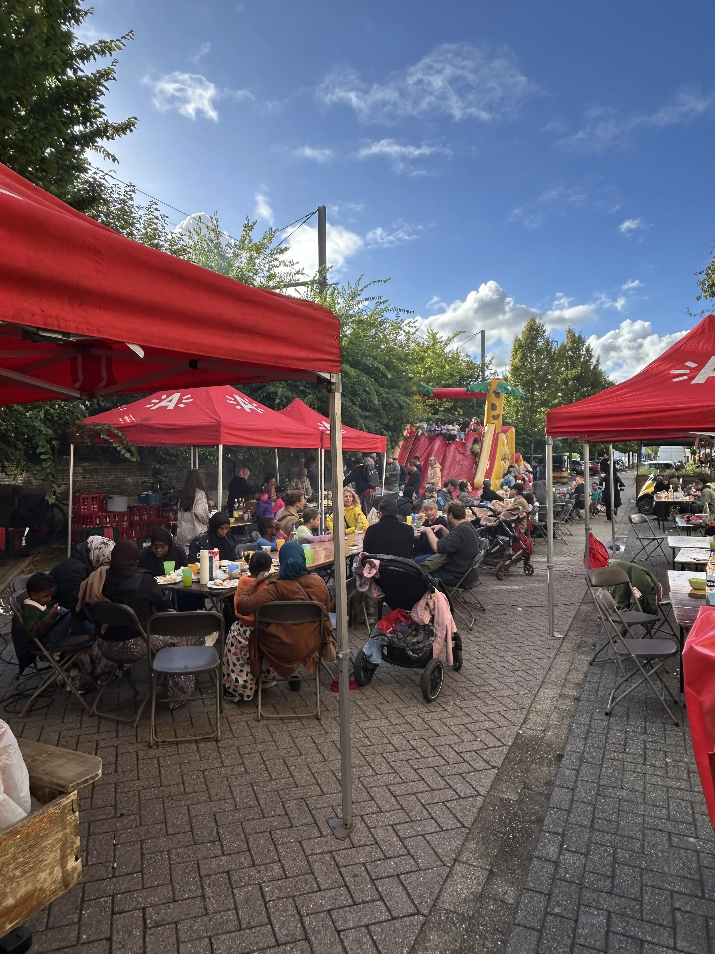 Gezellig buitenfeest met mensen die onder rode tenten eten en drinken, met een speeltoestel op de achtergrond, en een heldere blauwe lucht met enkele wolken.