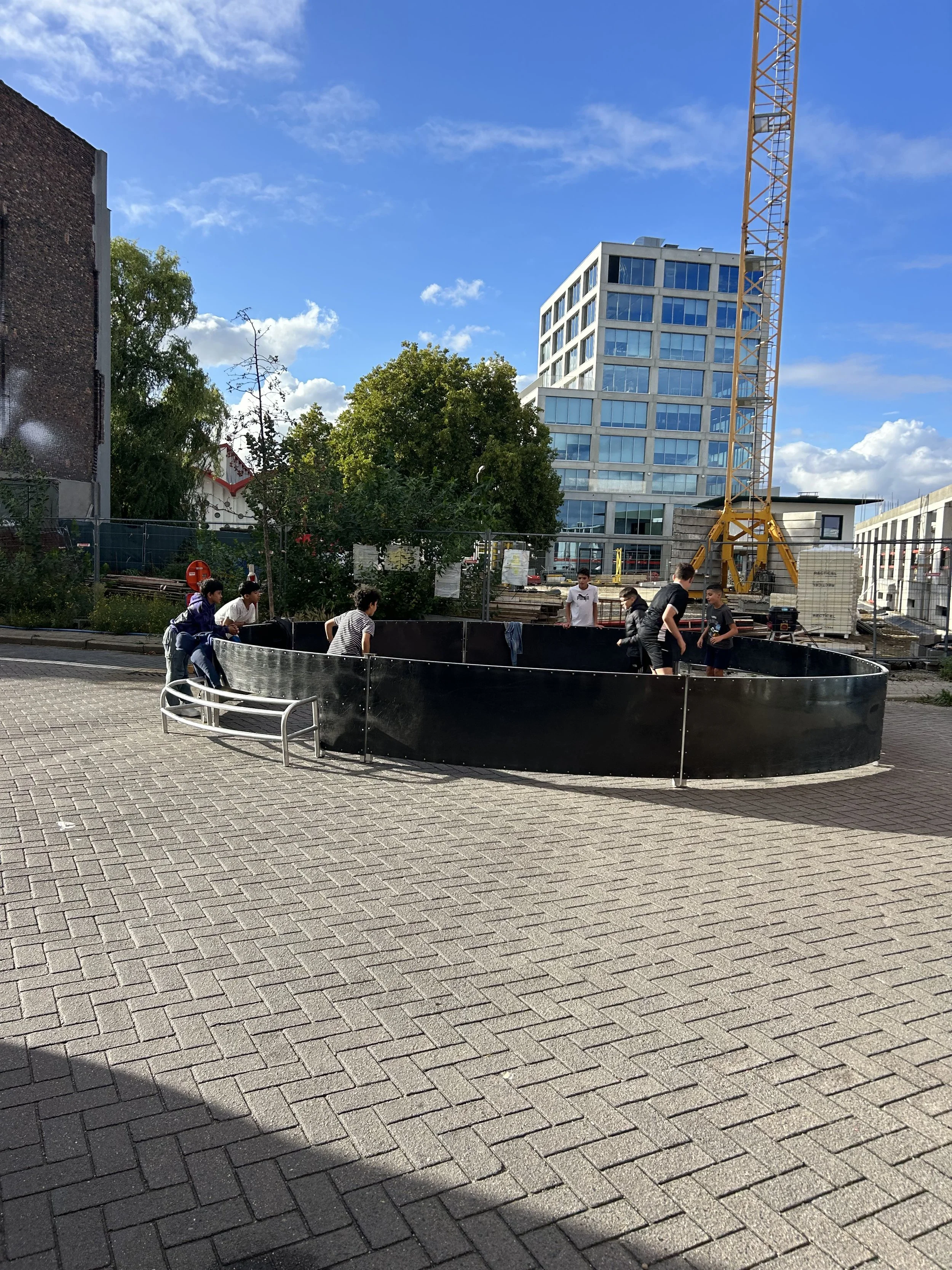 Groep kinderen speelt in een klein attractiecarrousel op een stadsplein met bouwprojecten en moderne gebouwen op de achtergrond. Situatie onder een blauwe hemel met wolken.
