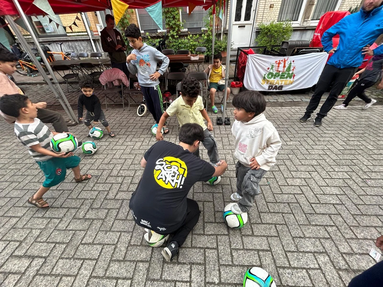 Kinderen die oefenen met voetballen onder begeleiding tijdens een buiten evenement, met een tent en groep volwassenen op de achtergrond.