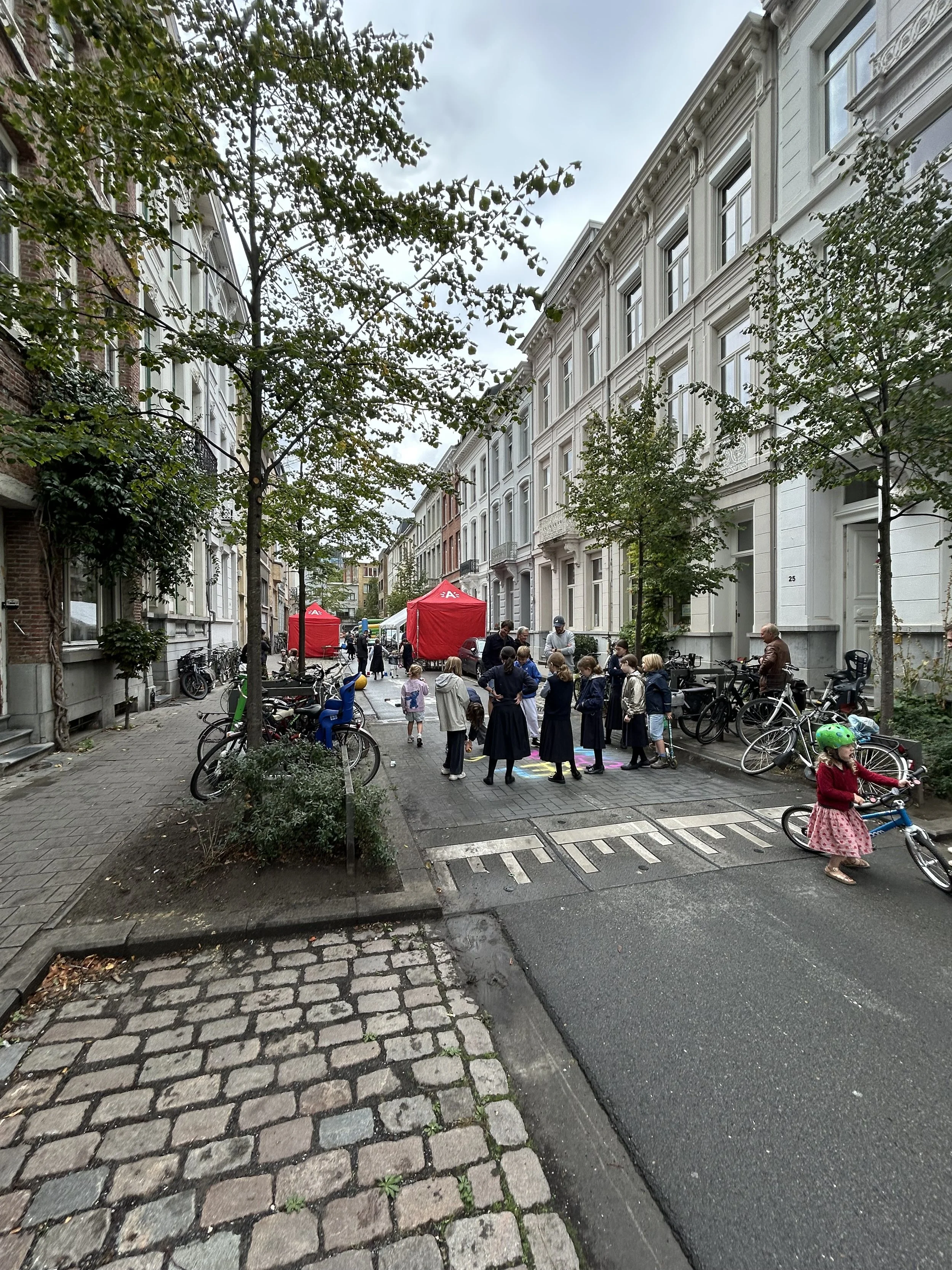 Kinder en volwassenen verzamelen zich op een straat tussen witte en gekleurde historische gebouwen, met rode tenten op de achtergrond en fietsen geparkeerd langs de zijkant.