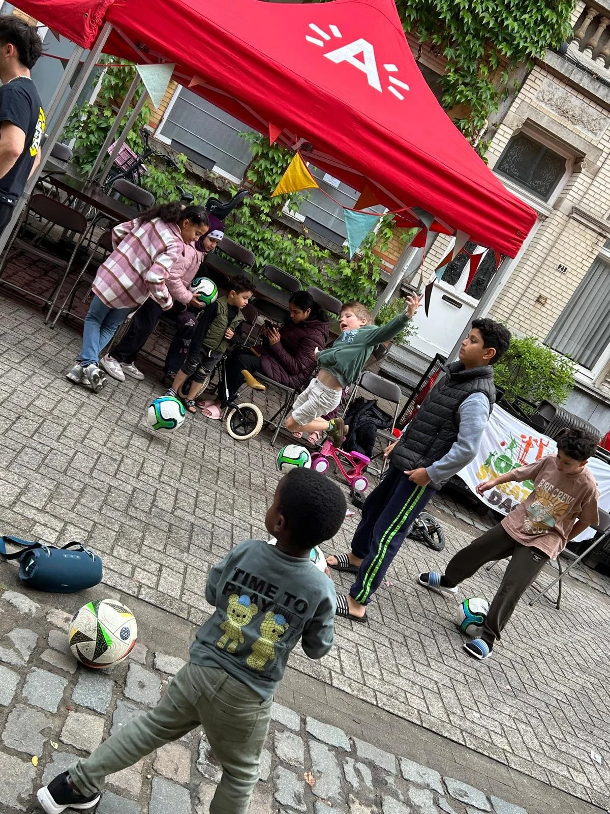 Kinderen en volwassenen bij een street soccer evenement onder een rode tent met logo, spelend met voetbal en skates, in een straat met stenen en tegels.