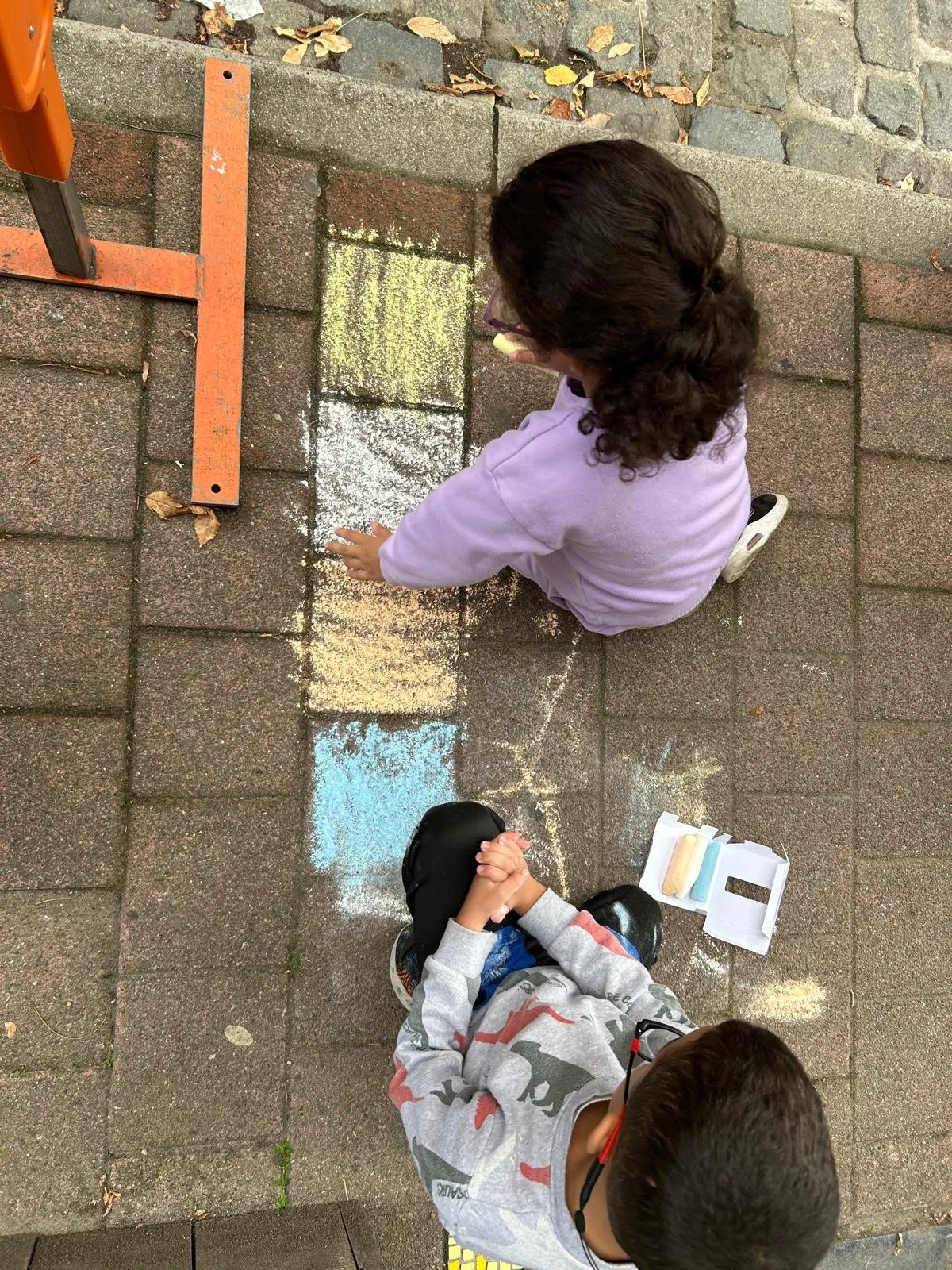 Twee kinderen die samen op straat met gekleurde krijtjes tekenen en de hand van elkaar vasthouden.
