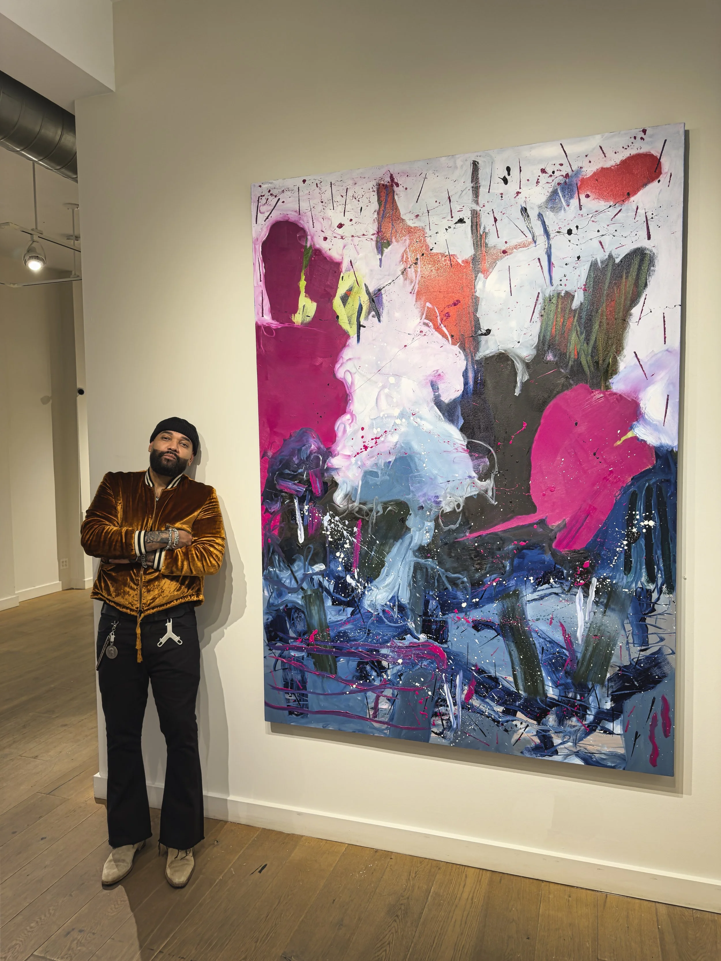 A man stands with arms crossed next to a large abstract colorful painting in an art gallery.