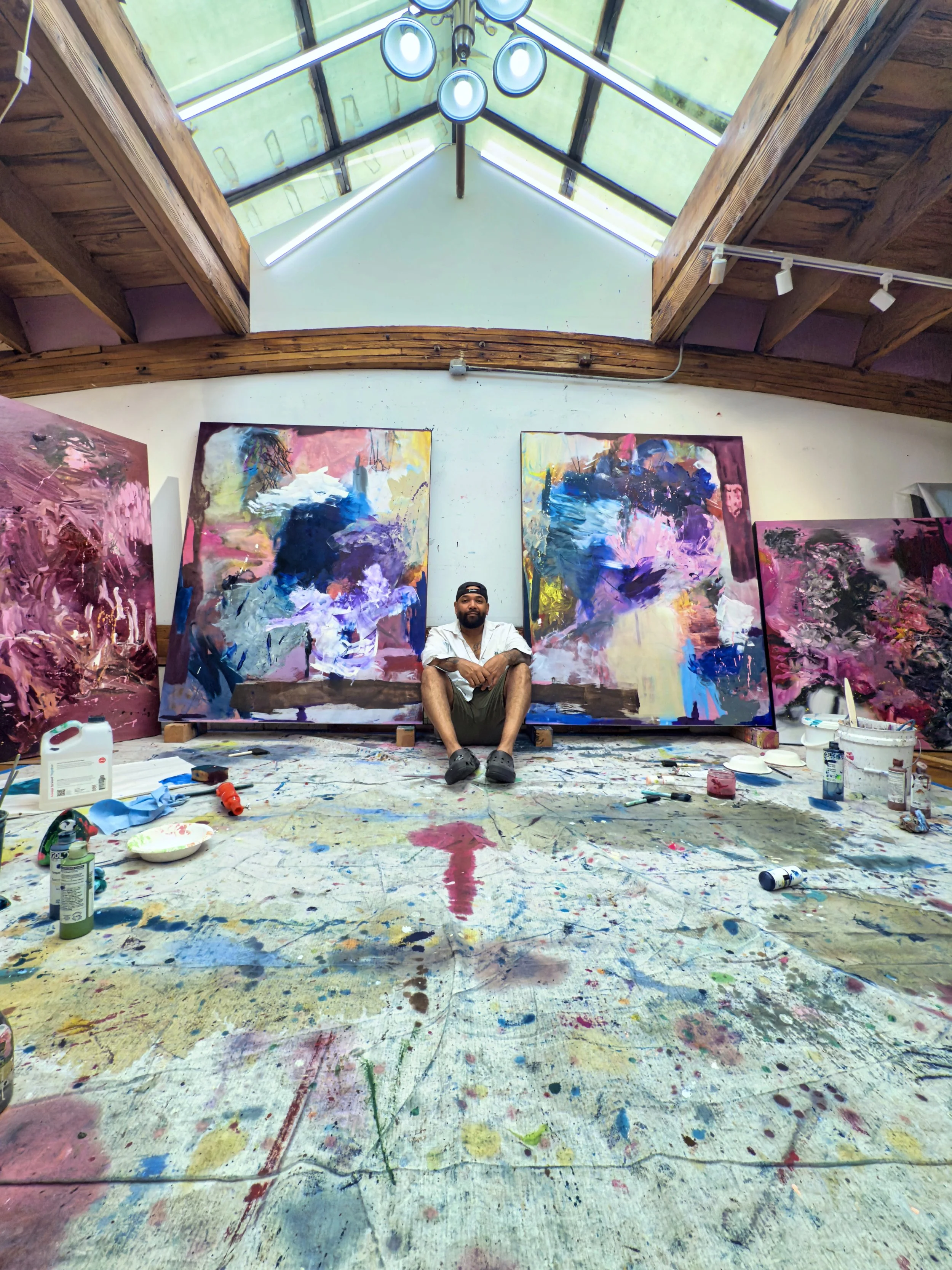 An artist sitting in an art studio surrounded by colorful abstract paintings, with paint supplies scattered on the floor and a large skylight overhead.