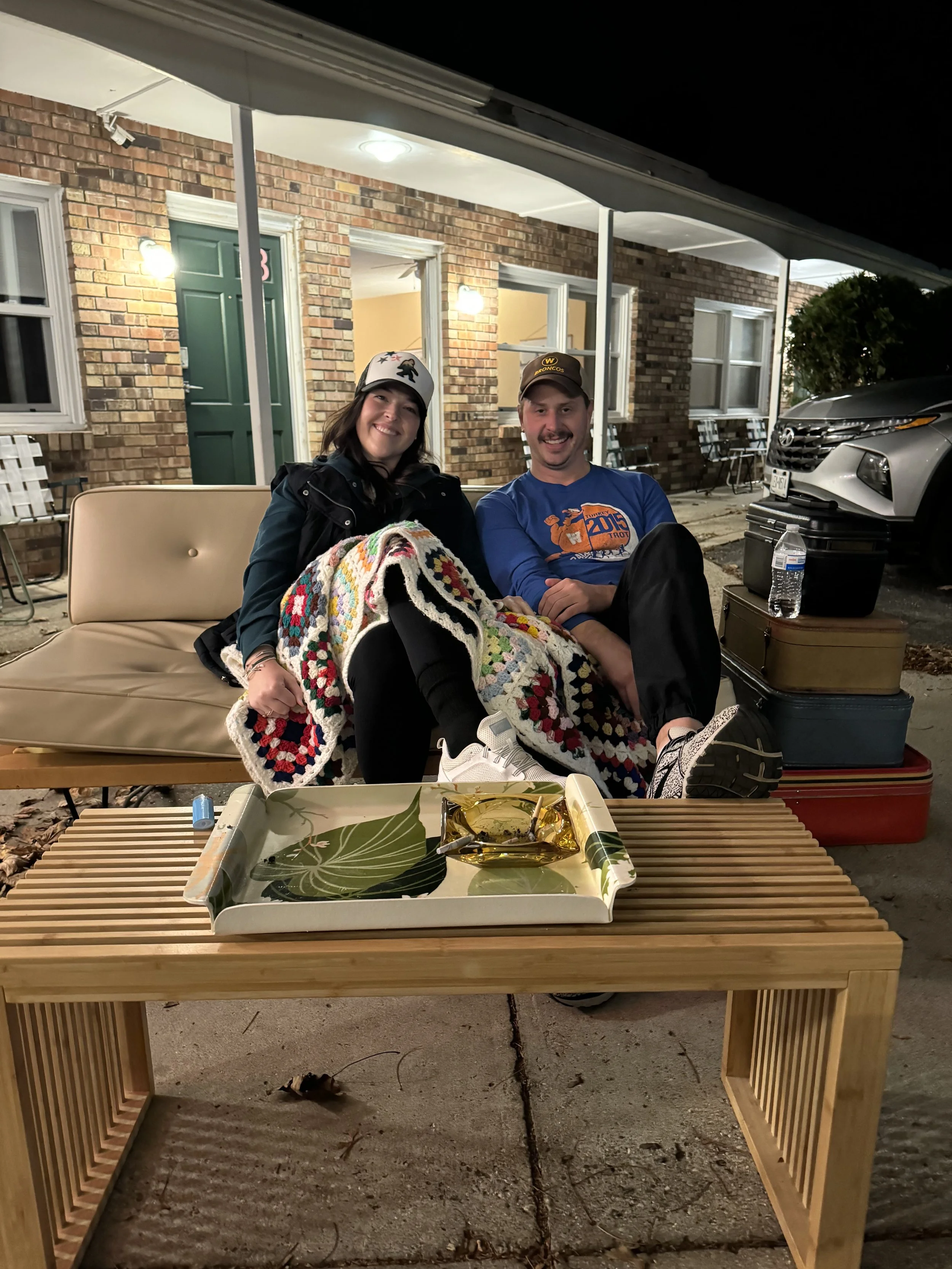 A man and a woman sitting on a couch outside at night, smiling. The woman is wrapped in a colorful crocheted blanket and the man is wearing a blue shirt and a cap. There are chairs and a car in the background, with a Tray of snacks and a bottle of water nearby.
