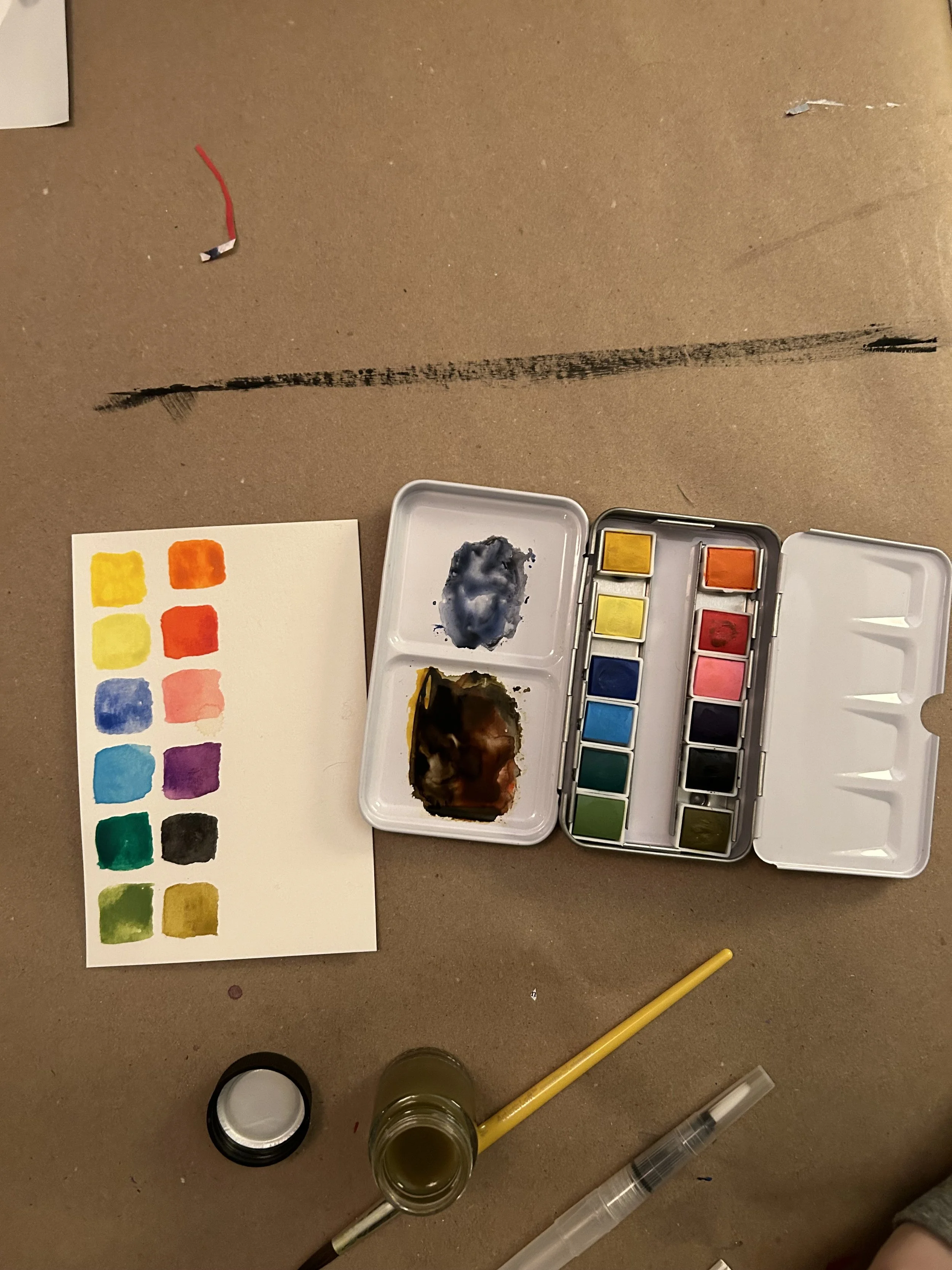 Paint palette with watercolor paints, a color swatch on paper, and various painting tools on a brown paper surface.